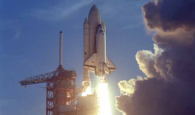 Backed by a blue sky, a space shuttle rises from the launchpad. White-hot flames emerge from the twin rockets powering the takeoff.