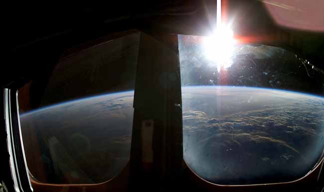 Peering through a window portal of the spacecraft, the view captures the curve of the top of Earth and above it, a brilliant sun.