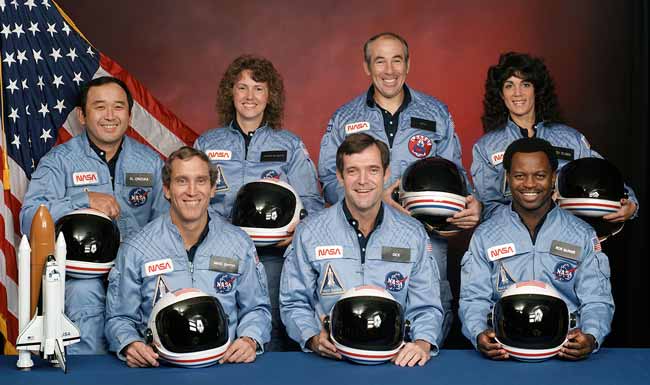 A studio portrait of a flight crew of astronauts. Three men in front are seated shoulder to shoulder. Their white helmets with black-tinted visors lie on a table top in front of the men. Four more astronauts - two men and two women - stand behind the trio. All are wearing pale blue flight suits. At left on the tabletop is a scale model of their spacecraft.