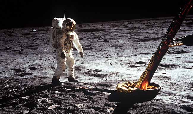 A fully-suited astronaut stands on the gray surface of the moon. The astronaut's face visor reflects what he sees. At right is the landing leg of his spacecraft.